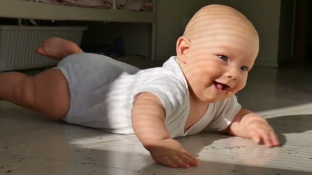 Um lindo bebê de meio ano está no chão e sorri alegremente. A criança bate um riacho no chão e empurra vigorosamente as pernas
 - Filmagem, Vídeo