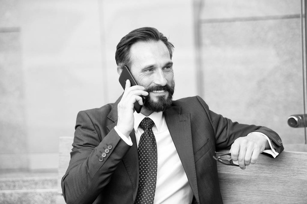 Over the phone. Portrait of modern businessman talking on smart-phone while sitting on bench outdoors. bearded man holding glasses in hand, talking on phone and smiling. - Photo, image