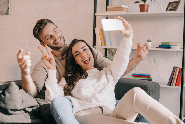 boldog fiatal pár selfie smartphone és béke gesztus a kanapén, otthon - Fotó, kép