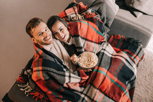 high angle view of young couple relaxing on couch with bowl of popcorn and covering with plaid - Foto, Imagen