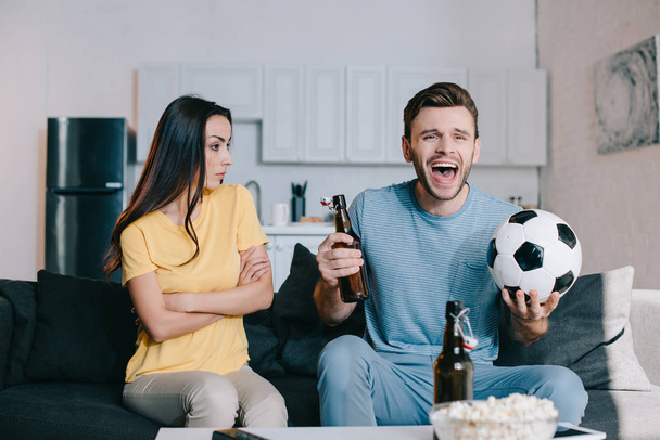 Młoda kobieta, patrząc na jej mąż krytycznie podczas oglądania meczu piłki nożnej i doping w domu - Zdjęcie, obraz