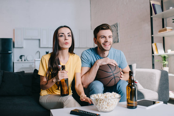 Happy νεαρό ζευγάρι, βλέποντας παιχνίδι μπάσκετ στην τηλεόραση στο σπίτι - Φωτογραφία, εικόνα