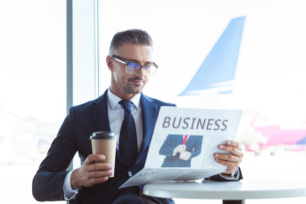 adulto guapo hombre de negocios leyendo periódico y la celebración de café para ir en el aeropuerto
 - Foto, Imagen