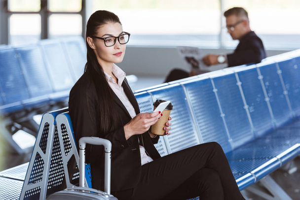 young businesswoman in glasses sitting with smartphone and coffee to go at airport - Zdjęcie, obraz