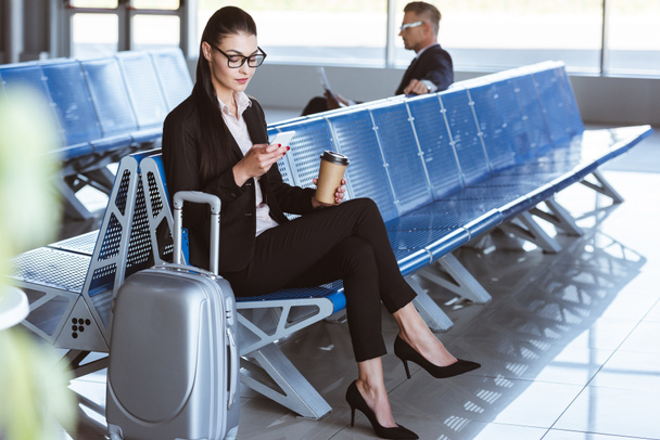 jonge zakenvrouw in glazen met behulp van smartphone in de vertrekhal op luchthaven - Foto, afbeelding