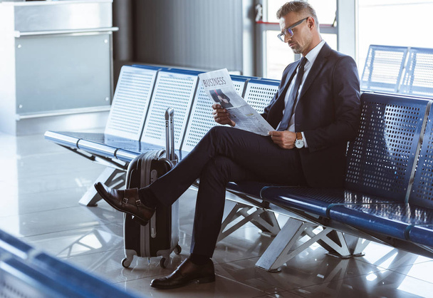 adult businessman sitting at departure lounge at airport and reading newspaper - Foto, Imagen