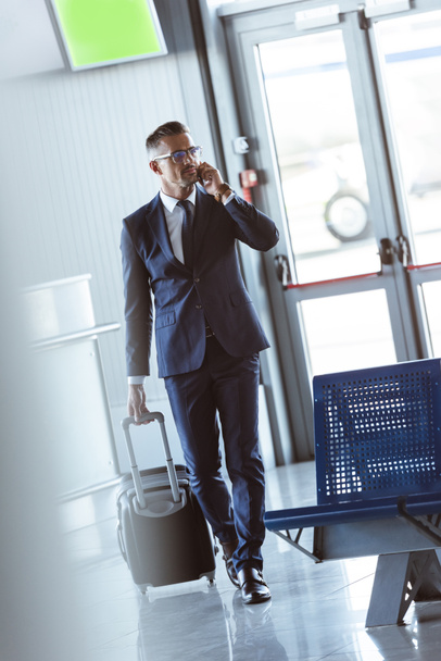 взрослый привлекательный бизнесмен со смартфоном и багажной прогулкой в аэропорту
 - Фото, изображение