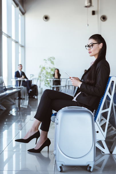 jonge zakenvrouw in glazen met smartphone zitten bij vertreklounge op luchthaven - Foto, afbeelding