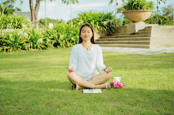 Young asian woman sitting with legs cross on the grass in a public park and meditating. Outdoor lifestyle photography. - Photo, Image