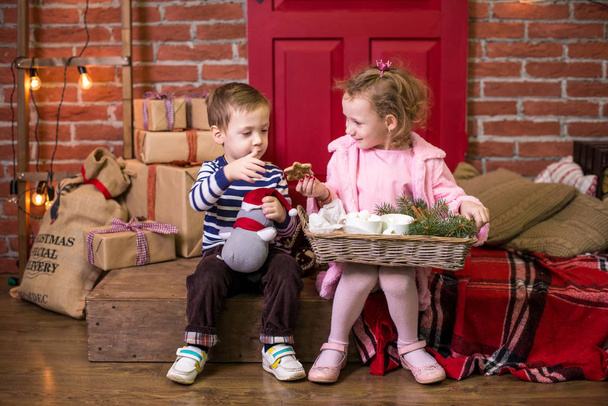 Due bambini piccoli, fratello e sorella, sono seduti sullo sfondo degli interni natalizi e dei regali. in possesso di un vassoio con dolci
. - Foto, immagini
