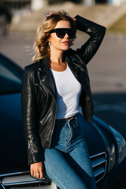 Portrait of young blonde woman in sunglasses standing next to black car - 写真・画像