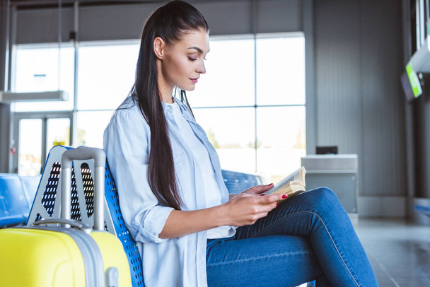 Beautiful woman with yellow luggage reading a book in the airport - Fotoğraf, Görsel