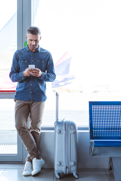 Elegante hombre de pie y mensajes de texto en el aeropuerto
 - Foto, imagen