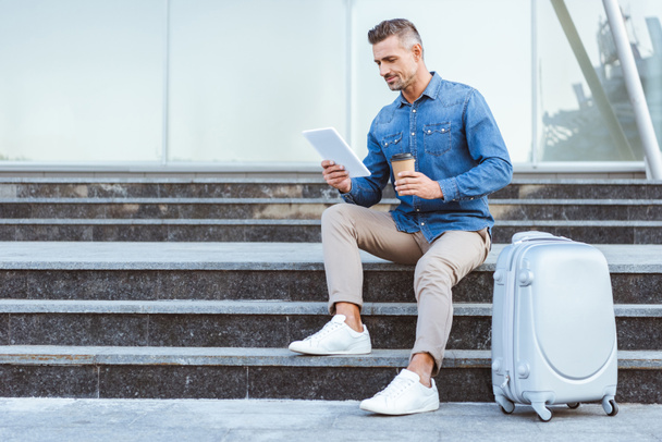 Handsome adult man with a coffee to go sitting on the staircase with luggage and using digital tablet - Fotografie, Obrázek