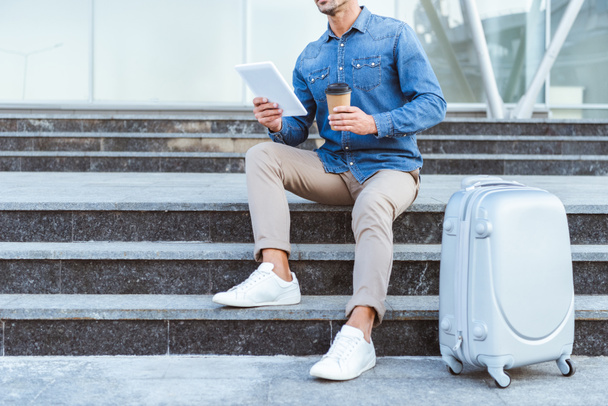 Man sitting on the staircase with luggage, holding digital tablet and coffee to go - Φωτογραφία, εικόνα