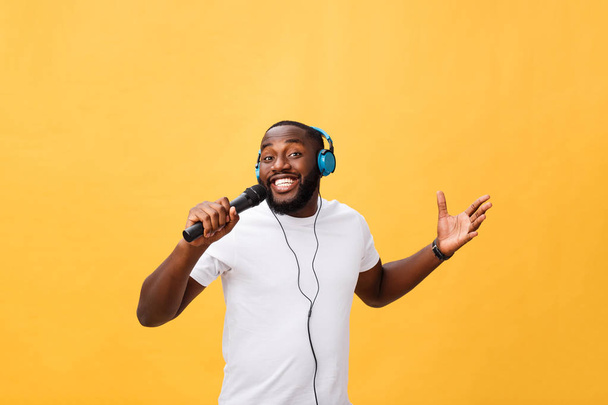 Portrait of cheerful positive chic handsome african man holding microphone and having headphones on head listening music singing song enjoying weekend vacation isolated on yellow background. - Фото, зображення
