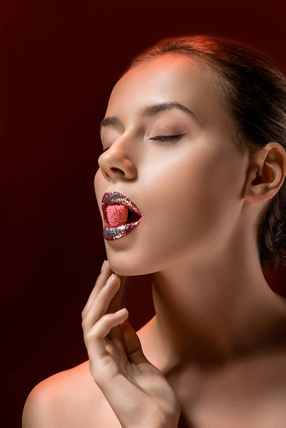 young woman with shiny lips and closed eyes holding pink candy in mouth on burgundy background - Фото, изображение
