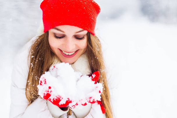 A menina segura a neve em suas mãos e olha para ele. belo sorriso e maquiagem para a menina em uma jaqueta de inverno e acessórios quentes
 - Foto, Imagem