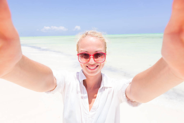 Młoda kobieta ubrana w białą plażę tunika biorąc selfie na tropikalnej, piaszczystej plaży. - Zdjęcie, obraz