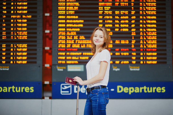 Young woman in international airport with luggage and passport near flight information display - Photo, Image
