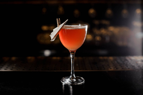 Glass of a delicious cocktail decorated with a paper plane standing on the barstand on the blurred dark and yellow light background - 写真・画像