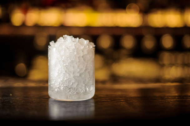 Cylindrical cocktail glass of crushed ice standing on the wooden bar counter on the blurred dark and light background - Fotografie, Obrázek