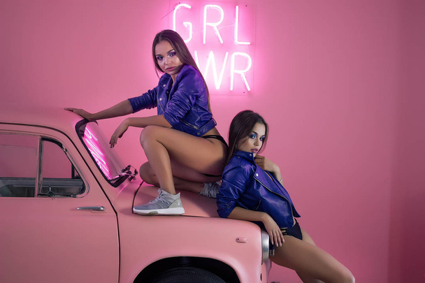 νέοι σέξι κορίτσια σε μπλε δερμάτινο μπουφάν, μαύρα εσώρουχα και χρώματα σε ροζ φόντο, μία κοπέλα κάθεται πάνω στο καπό του αυτοκινήτου ροζ, το δεύτερο στέκεται δίπλα της για ένα ροζ μεταλλικό κουτί - Φωτογραφία, εικόνα