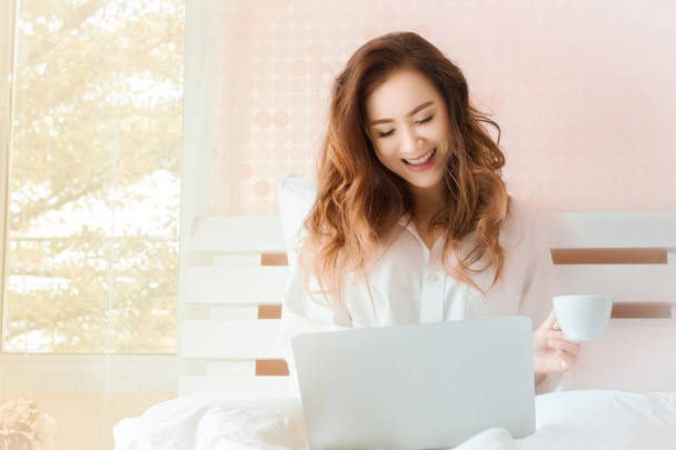 Young beautiful woman smiling and holding cup of coffee and used laptop on the bed with happiness, light warm in the morning and copy space - 写真・画像