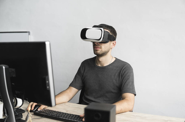 Young man wearing virtual reality goggles headset, vr box and sitting in the office against computer monitor. Connection, technology, new generation, progress concept. - Foto, imagen