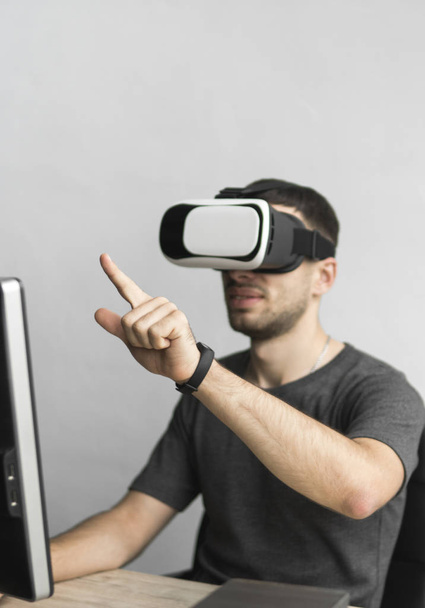 Young man wearing virtual reality goggles headset and sitting in the office against computer. Connection, technology, new generation. Man trying to touch objects or control VR with a hand. - Photo, Image