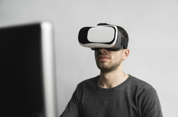 Young man wearing virtual reality goggles headset, vr box and sitting in the office against computer monitor. Connection, technology, new generation, progress concept. - Foto, afbeelding