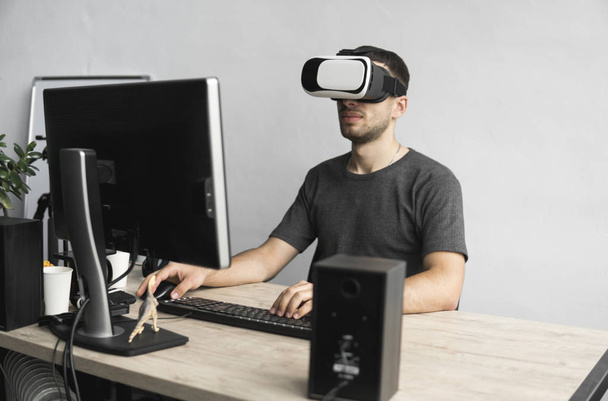 Young man wearing virtual reality goggles headset, vr box and sitting in the office against computer monitor. Connection, technology, new generation, progress concept. - Foto, imagen