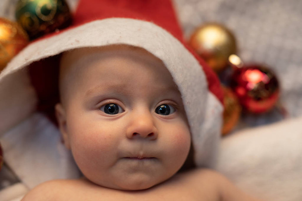 Joven bebé Santa sombrero con un rojo acostado en una cuna con adornos de Navidad rodeando su cabeza en un retrato de cerca de la cara
 - Foto, imagen