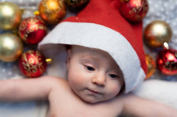 Cute tiny baby wearing a festive red Christmas Santa hat lying in its cot surrounded by red and gold baubles - Photo, Image