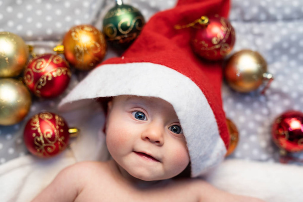 Adorable little baby dressed in a festive red Santa hat lying in its cot looking up at the camera with big eyes surrounded by Christmas decorations - Photo, Image