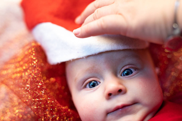Mère tenant un chapeau de Père Noël sur un mignon petit nouveau-né dans une crèche de Noël en regardant la caméra avec d'énormes yeux curieux
 - Photo, image