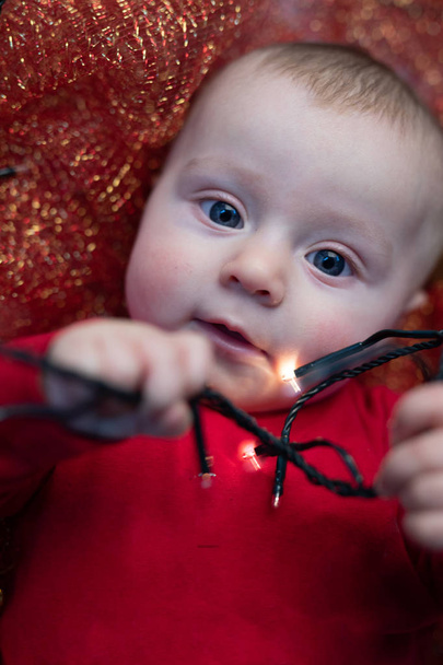 Adorável bebê de Natal brincando com luzes da festa enquanto vestindo uma roupa vermelha festiva Papai Noel enquanto deitado em seu berço
 - Foto, Imagem