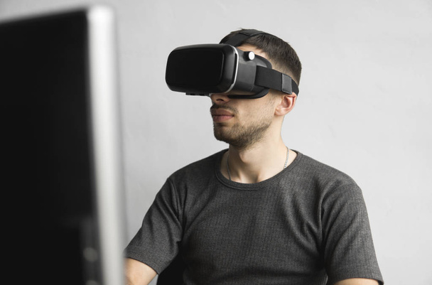Young man wearing virtual reality goggles headset, vr box and sitting in the office against computer monitor. Connection, technology, new generation, progress concept. - Fotoğraf, Görsel