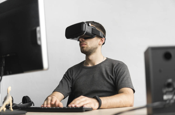 Young man wearing virtual reality goggles headset, vr box and sitting in the office against computer monitor. Connection, technology, new generation, progress concept. - Valokuva, kuva