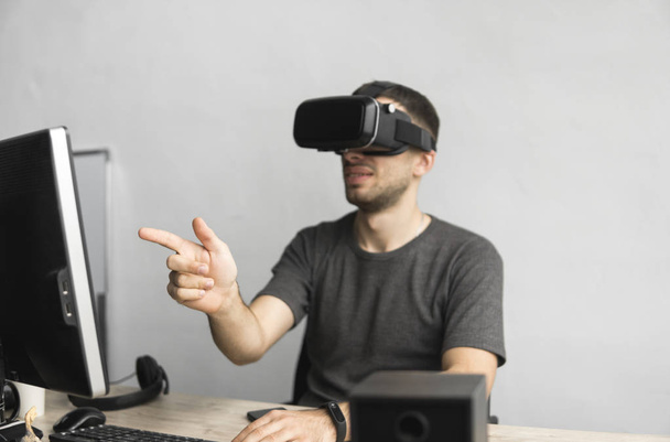 Young man wearing virtual reality goggles headset and sitting in the office against computer. Connection, technology, new generation. Man trying to touch objects or control VR with a hand. - Fotografie, Obrázek