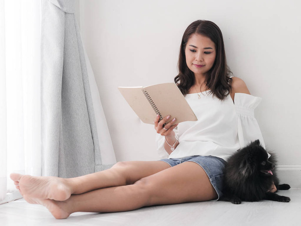 Happy Asian woman reading a book and sitting on floor with her black dog at home, lifestyle concept. - Фото, изображение