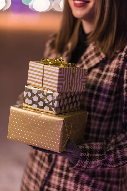 Jong meisje op de voorgrond staande op de kerstboom lichten bedrijf presenteert en geschenken en glimlachend in de winter alleen in het park. focus op de cadeautjes - Foto, afbeelding