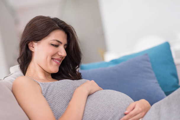 young happy pregnant woman keeping hand on belly while sitting on sofa at home - Foto, immagini