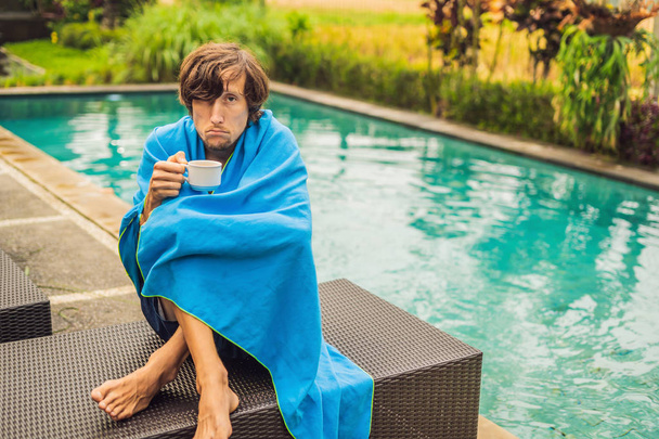 Sick man traveler. The man caught a cold on vacation, sits sad at the pool drinking tea and blows his nose into a napkin. His son is healthy and swimming in the pool. Travel insurance concept - Photo, Image