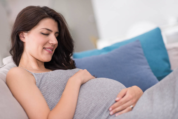 young happy pregnant woman keeping hand on belly while sitting on sofa at home - 写真・画像