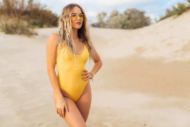 Sexy model in yellow swimsuit and sunglasses posing on the white sand beach. - Valokuva, kuva
