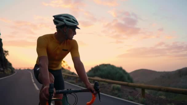 Een man op een fiets rijdt kijken naar de camera bij zonsondergang op een bergweg. Slow motion steadicam - Video