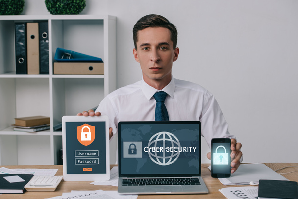 portrait of businessman showing laptop, tablet and smartphone with cyber security signs on screens at workplace in office - Photo, Image