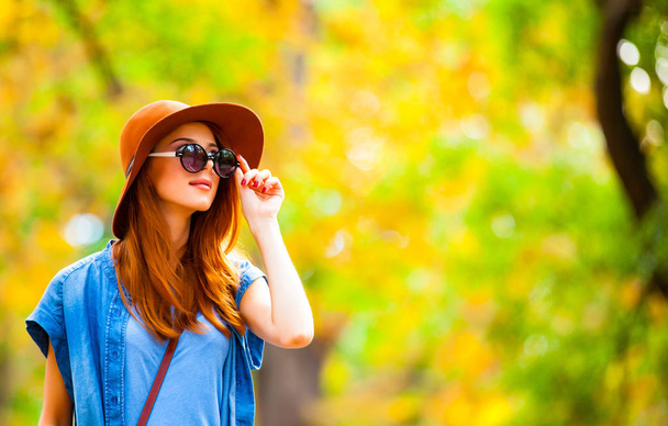 κοκκινομάλλα κορίτσι σε γυαλιά ηλίου και το καπέλο στο πάρκο φθινόπωρο. - Φωτογραφία, εικόνα