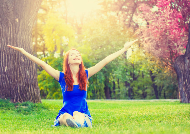 Happy redhead girl in blue dress sitting on green lawn. - Zdjęcie, obraz